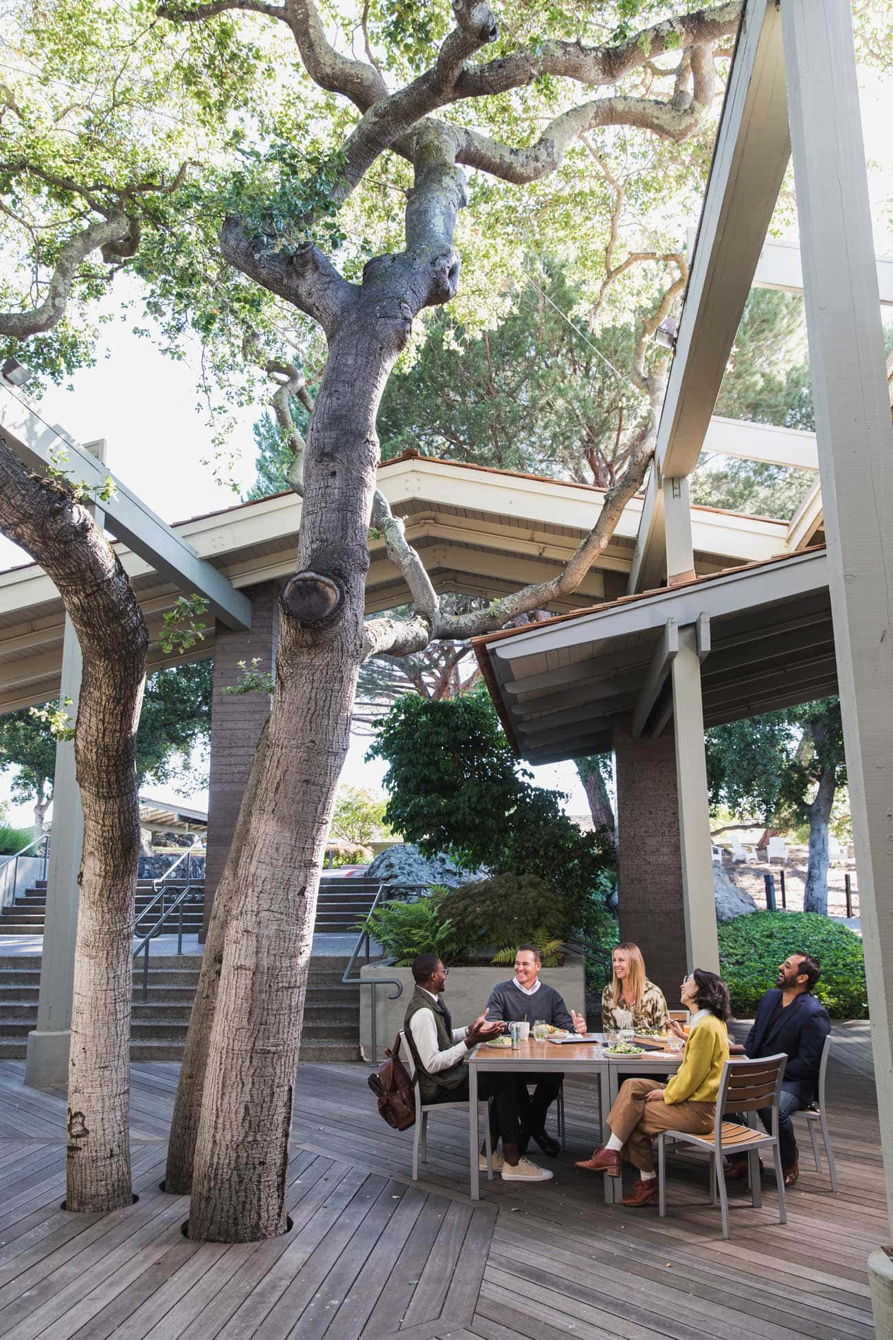 A group of colleagues sit together at a patio table surrounded by trees