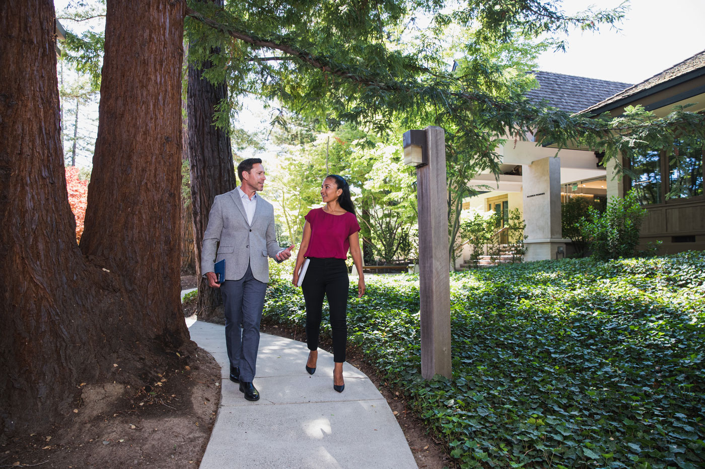 Two colleagues walk together on a landscaped sidewalk lined with trees